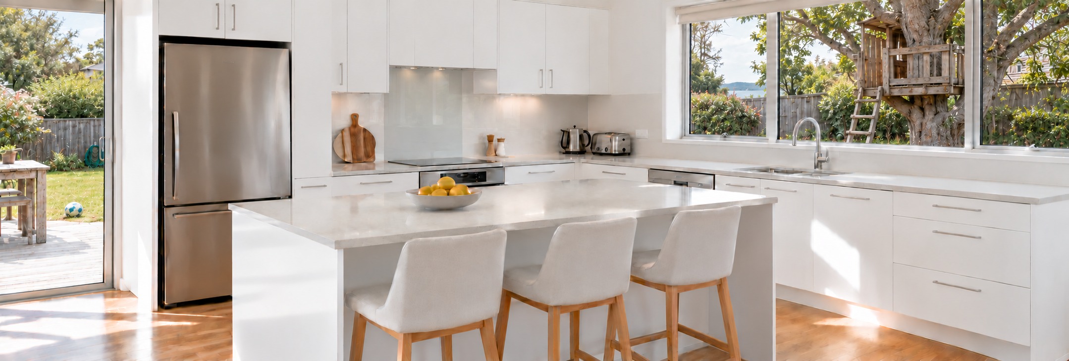 A bright white kitchen overlooking a family backyard with a timber treehouse